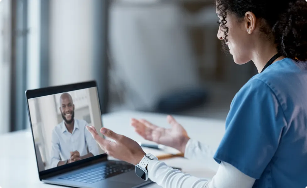Female healthcare worker in blue scrubs speaking during a telehealth call on a laptop.